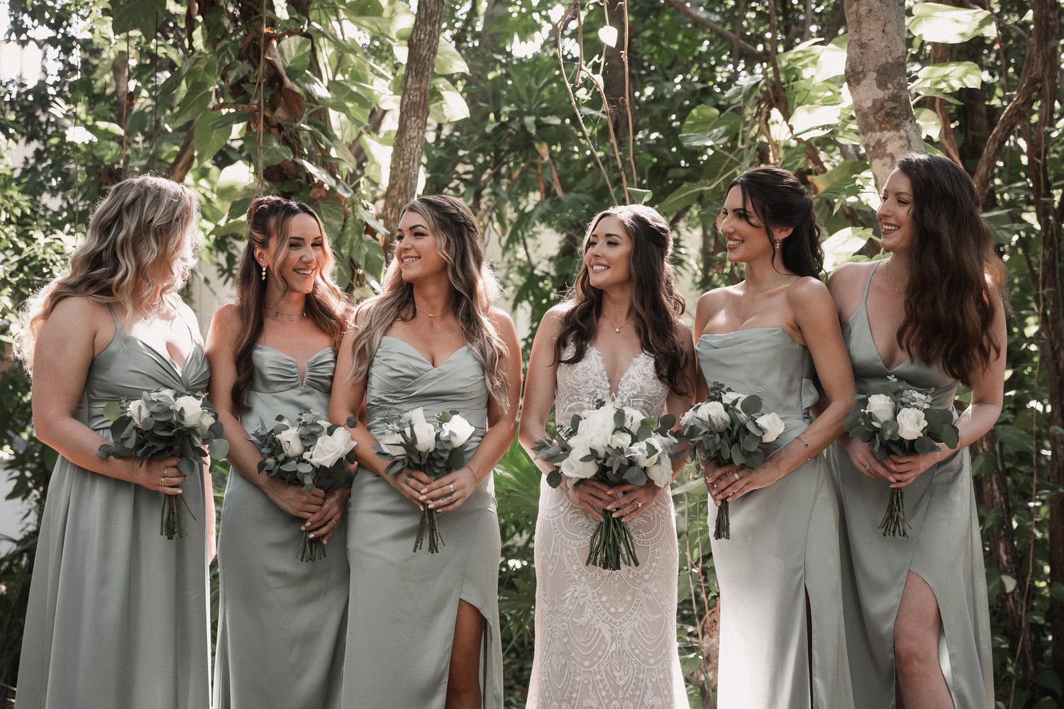 Bride and five bridesmaids in sage green dresses holding white bouquets laughing in tropical garden.