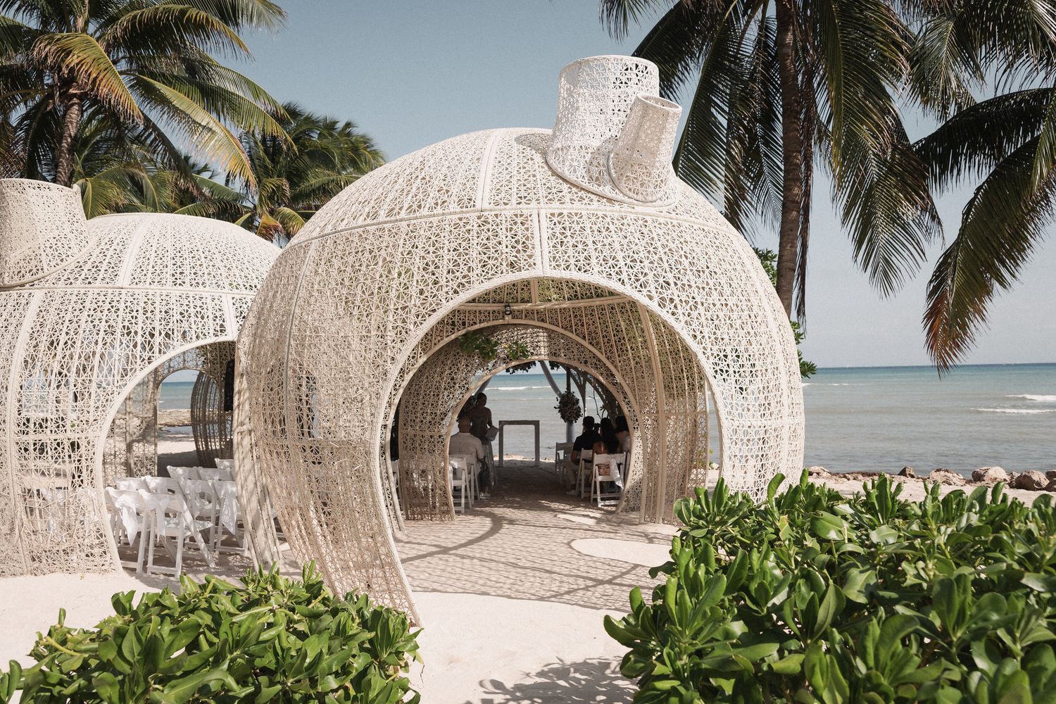White wicker dome-shaped beach cabanas on a tropical shoreline with palm trees and turquoise ocean views.