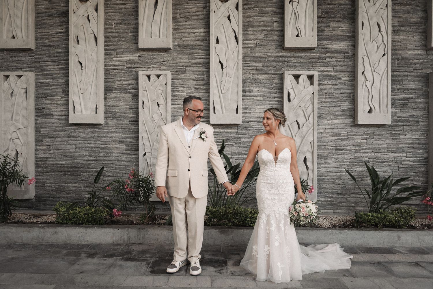 Newlyweds hold hands smiling before decorative stone wall, bride in lace mermaid gown, groom in cream suit.