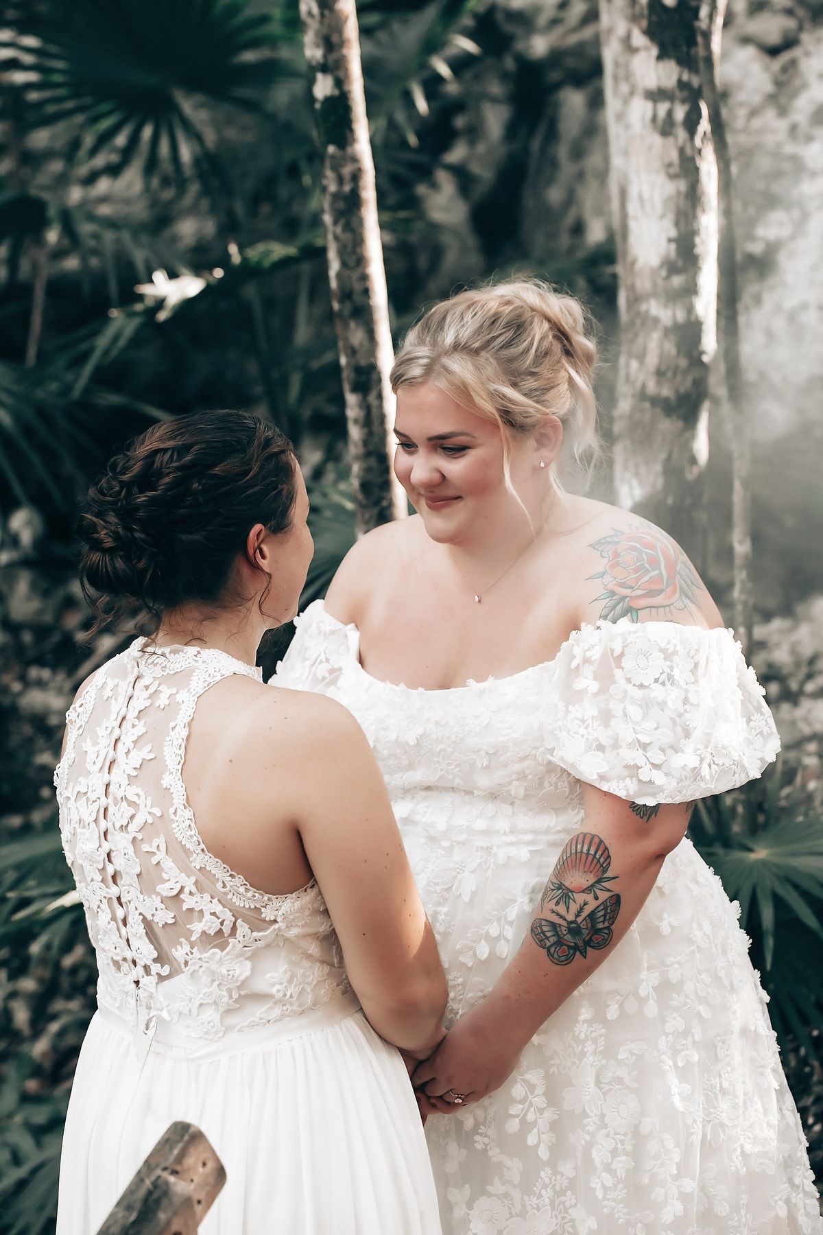 The image shows two women in white wedding dresses embracing each other in a lush, tropical setting.