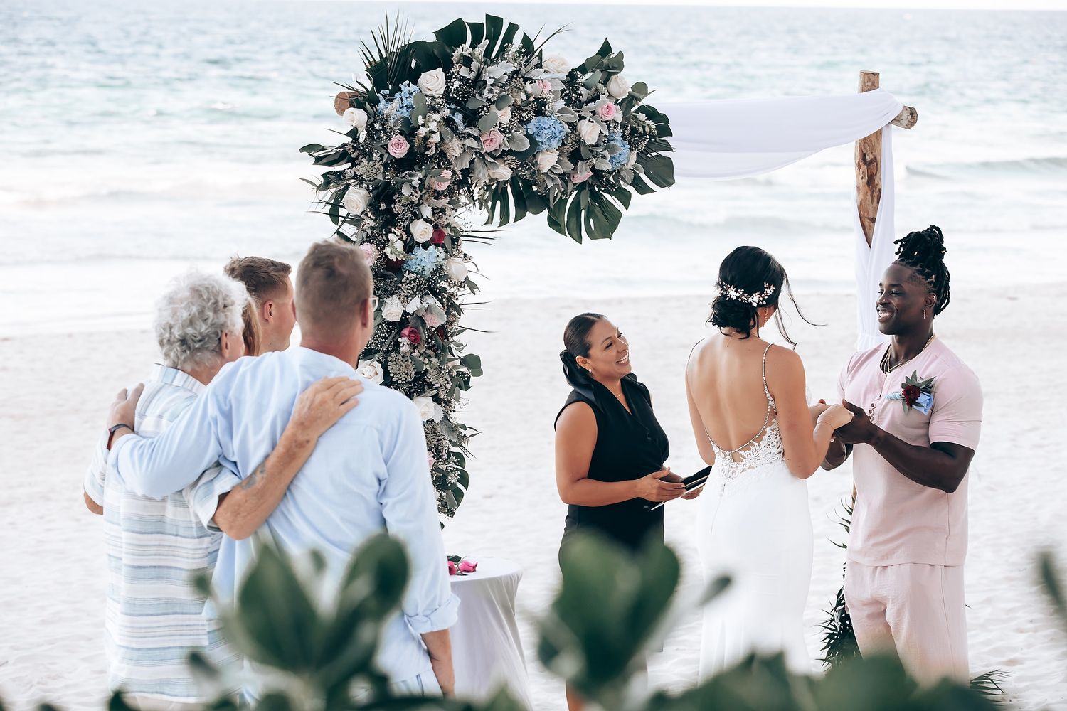 A wedding ceremony taking place on a beach, with a floral arch and guests in attendance.