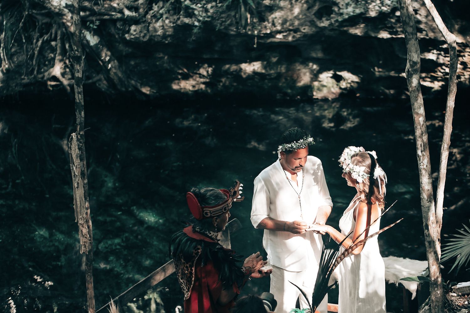 A couple exchanging vows in a tropical forest setting, with a person in traditional dress nearby.