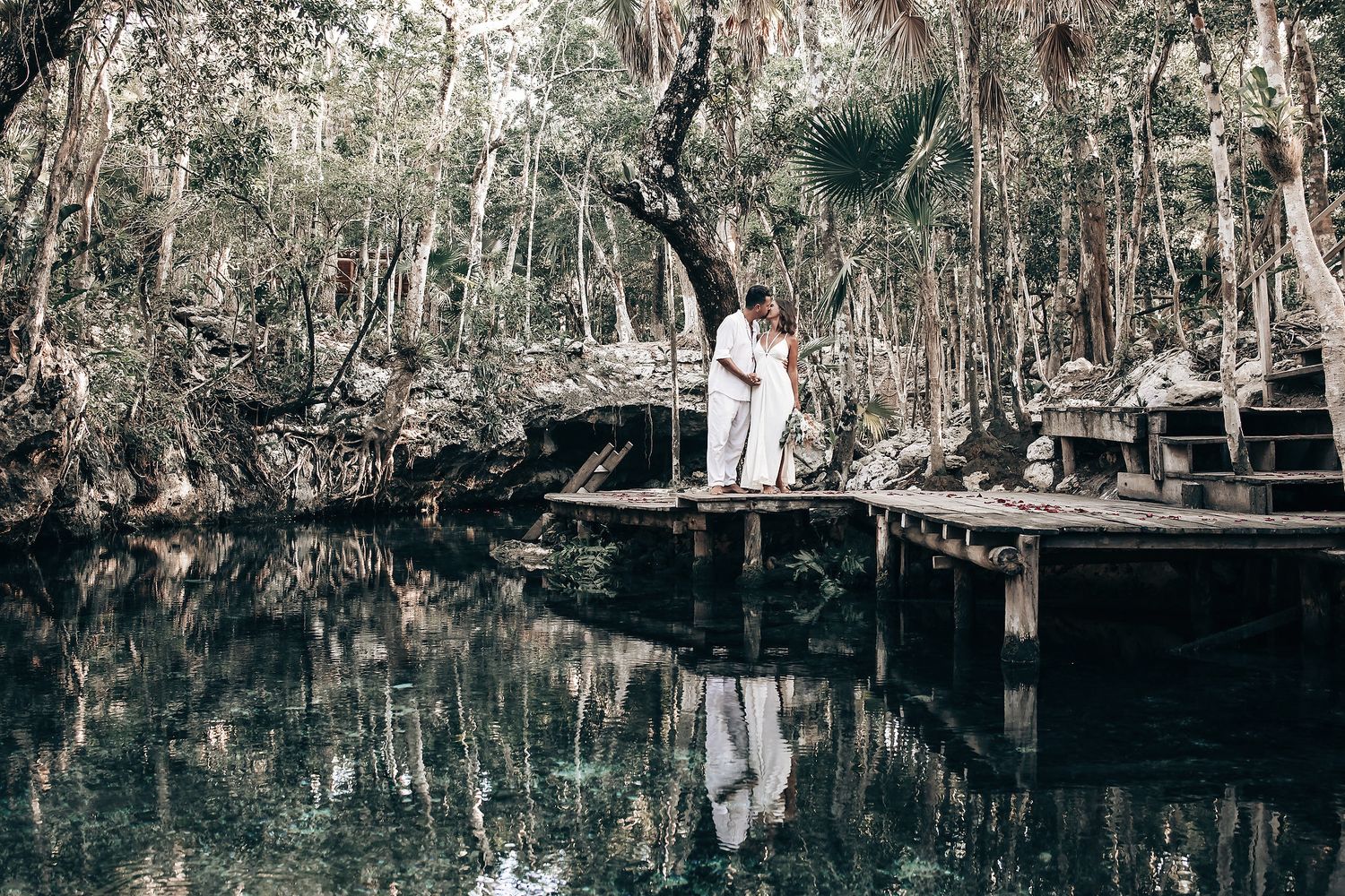 A couple stands together in a lush, forested landscape, surrounded by greenery and a body of water.