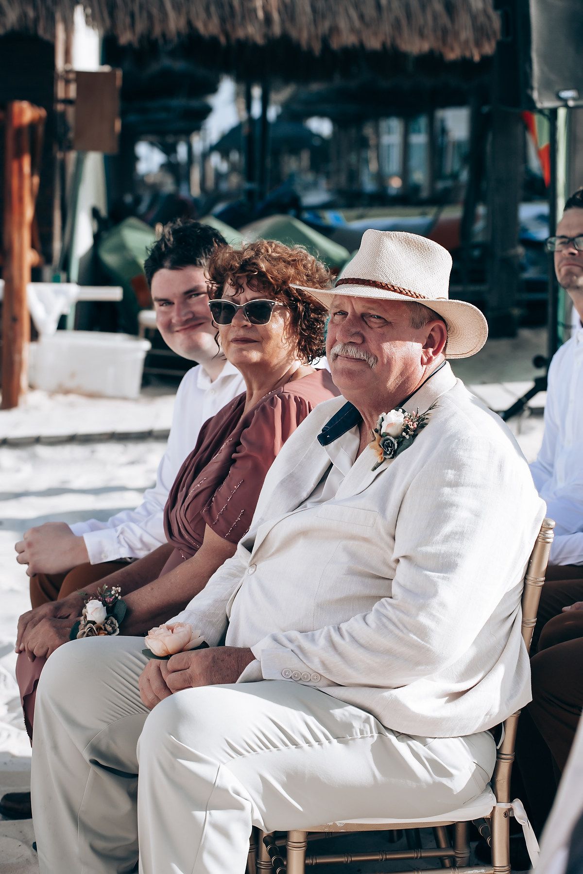 Wedding guests in white and casual attire seated in wooden chairs at outdoor venue.