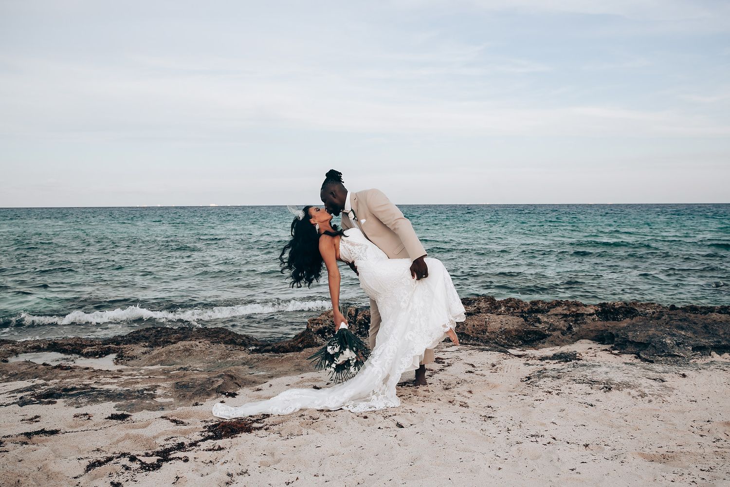 A romantic coastal wedding photoshoot with couple embracing on a rocky beach with ocean waves in the background.