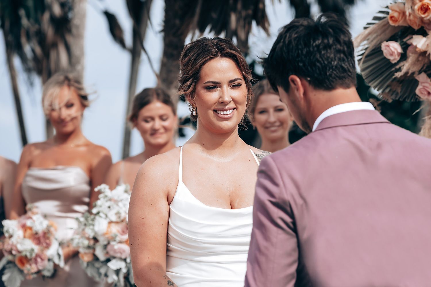 Wedding party members in neutral-toned dresses gather on a sunny beach with palm trees in the background.