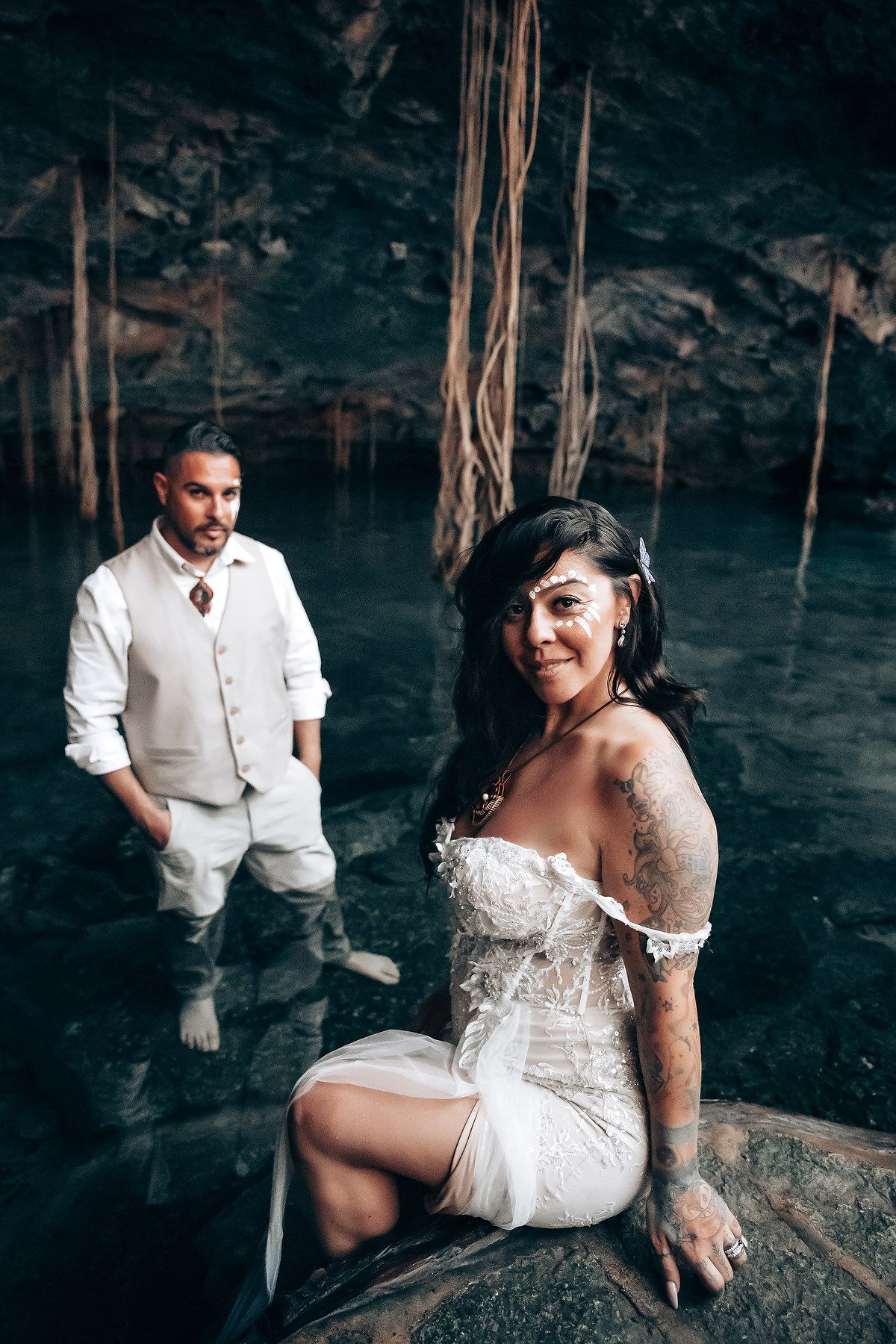 A couple poses together on rocky terrain inside a mystical cave with natural vine formations.