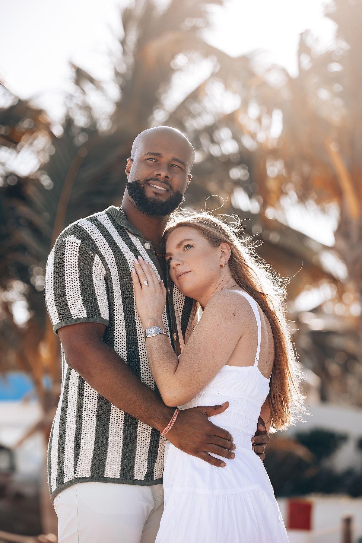 Interracial couple embracing warmly at golden hour with tropical palm trees glowing in background.