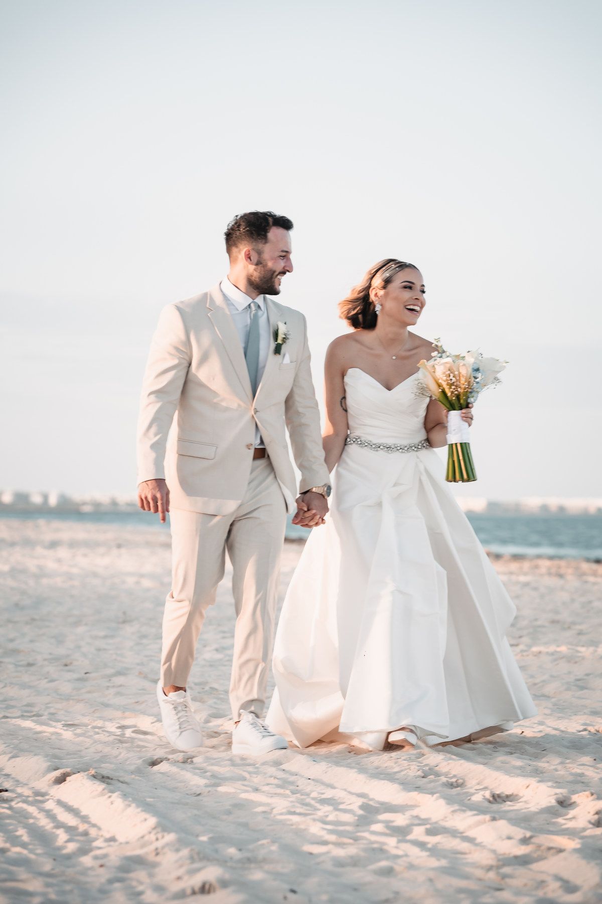 Bride and groom walk hand-in-hand on sandy beach at sunset in wedding attire.