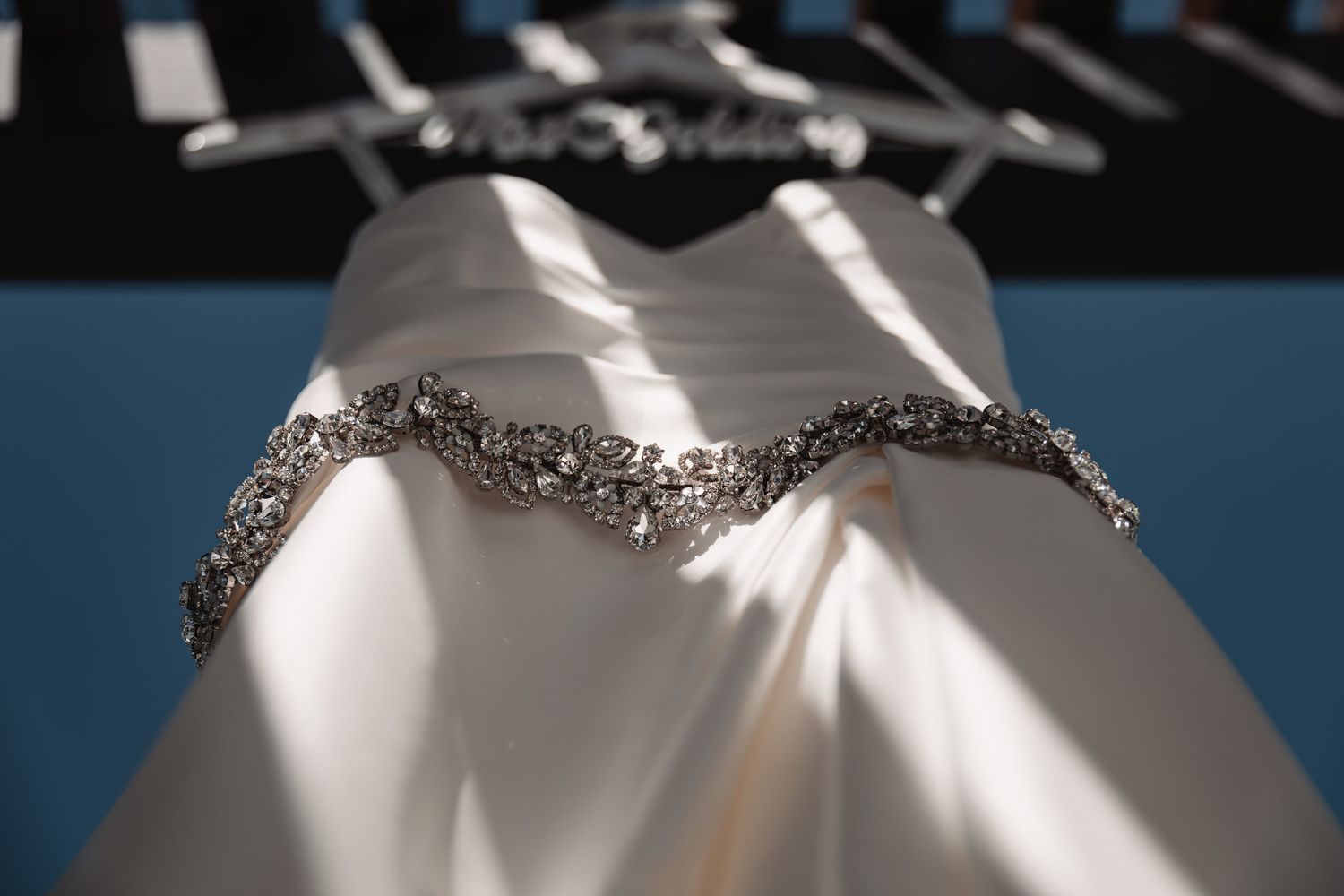 Elegant white wedding dress with intricate silver rhinestone beaded belt displayed on a hanger in soft natural light.