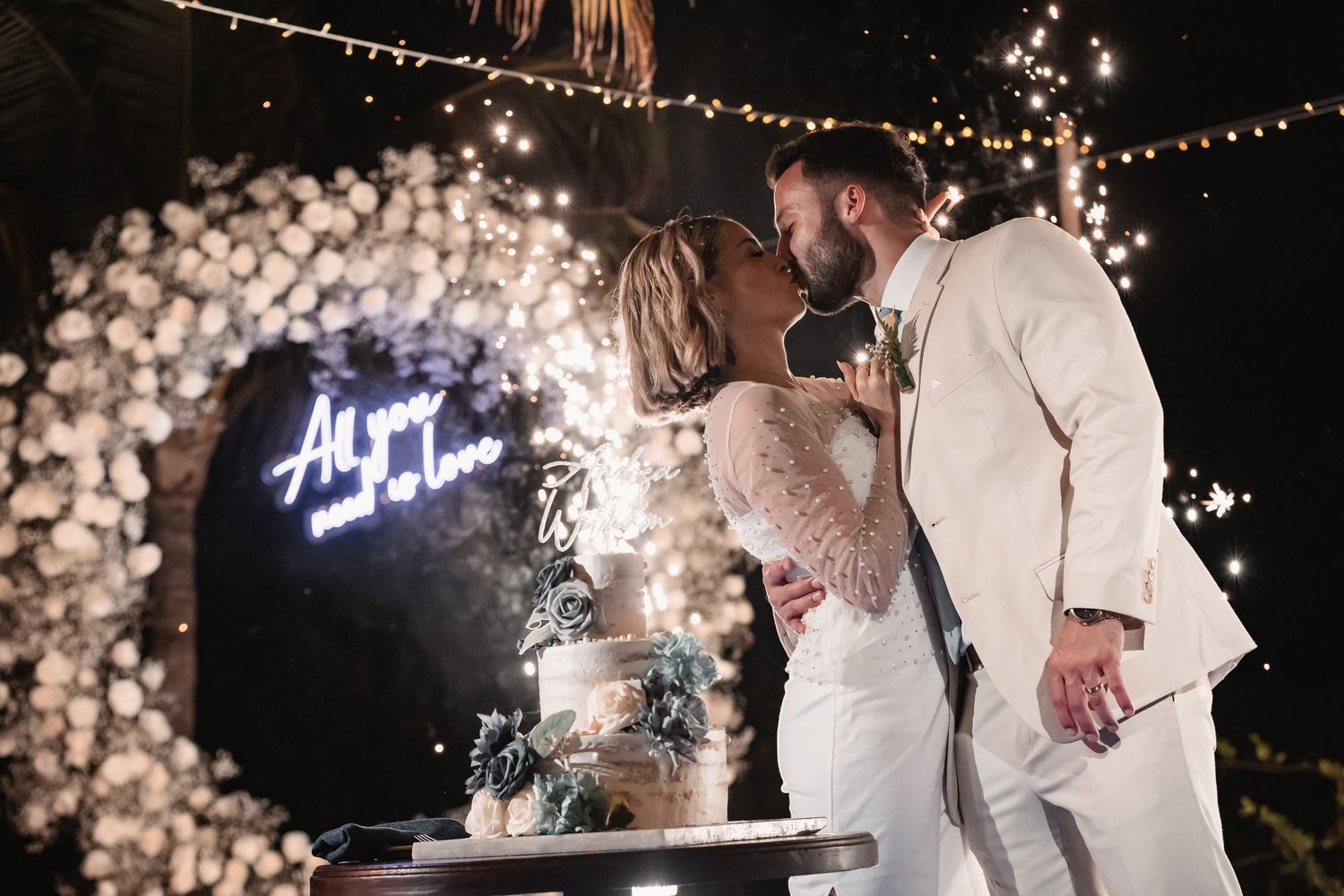Bride and groom kiss beside wedding cake under All You Need Is Love neon sign with sparklers.