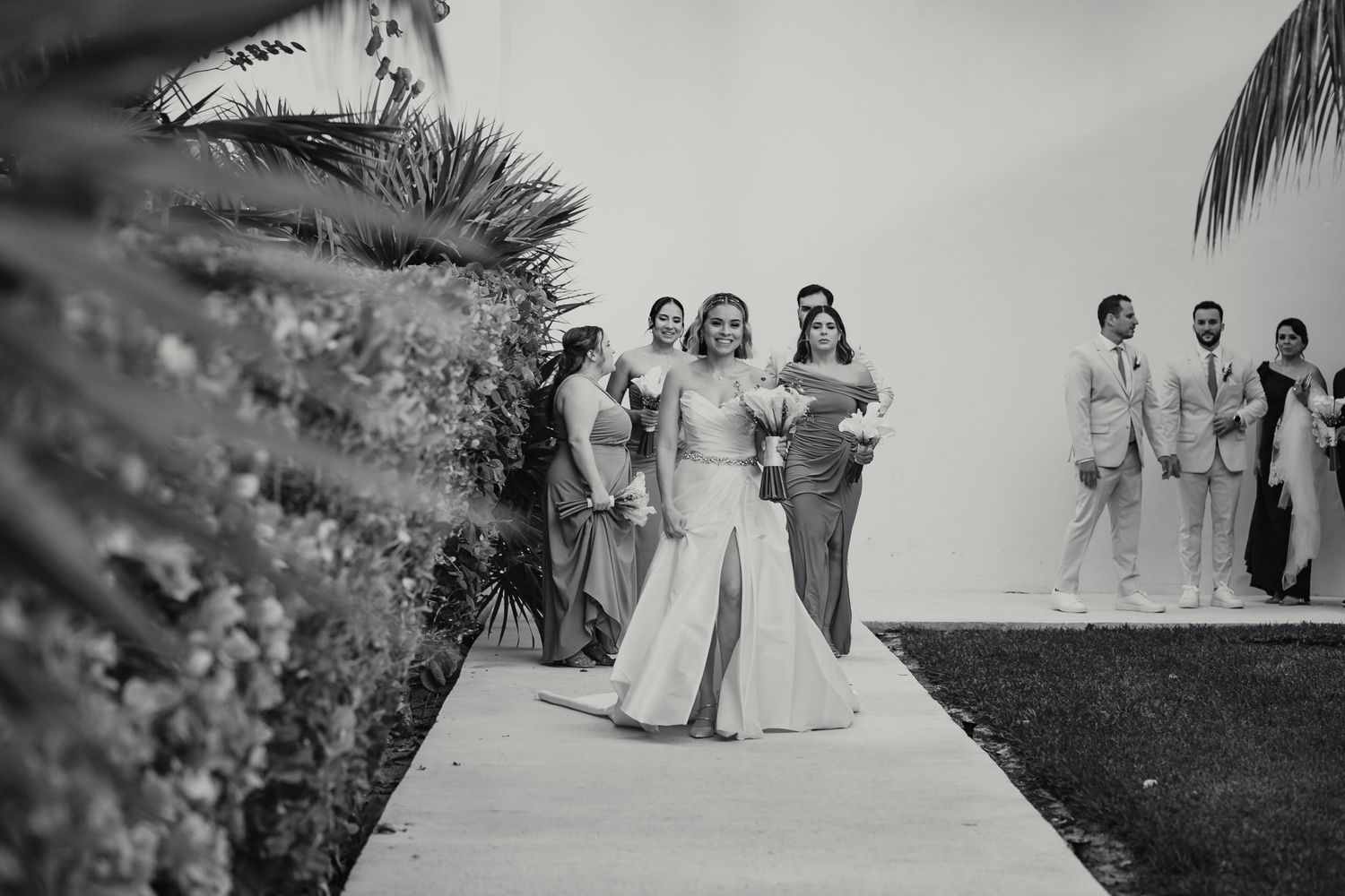 Black and white photo of bride walking down outdoor aisle with wedding party at tropical venue.