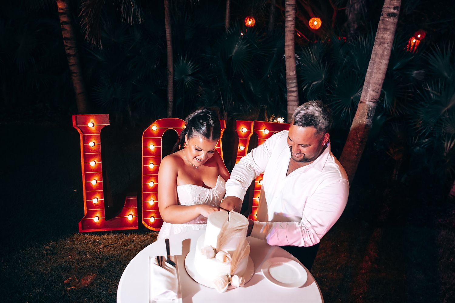 Couples dancing together at a nighttime outdoor wedding reception with red illuminated letters.