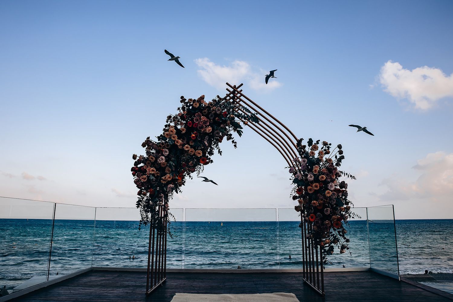Floral wedding arch with birds flying overhead against an ocean backdrop at sunset.
