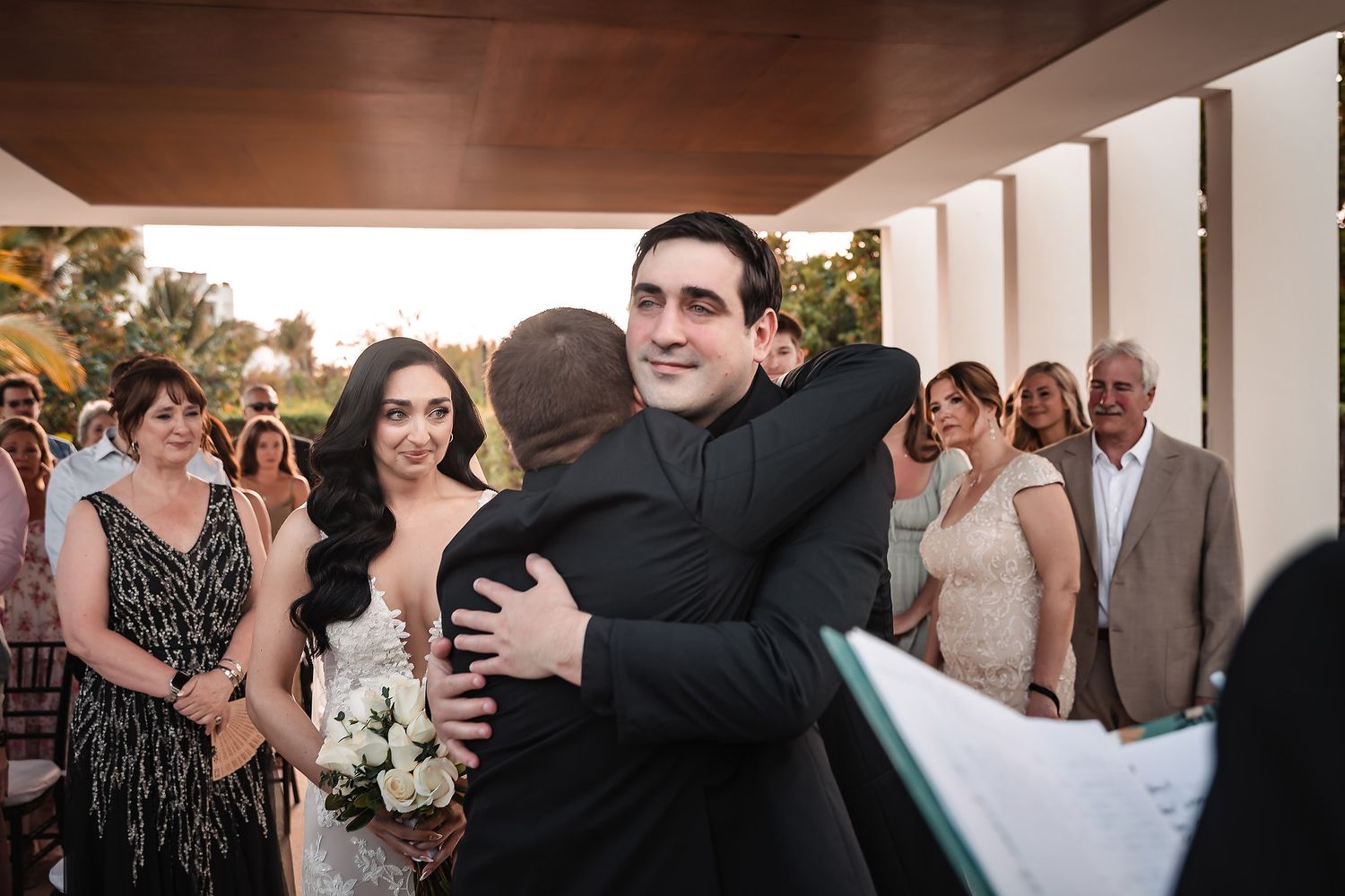 Groom hugging a guest at outdoor wedding ceremony while bride stands nearby holding white bouquet.