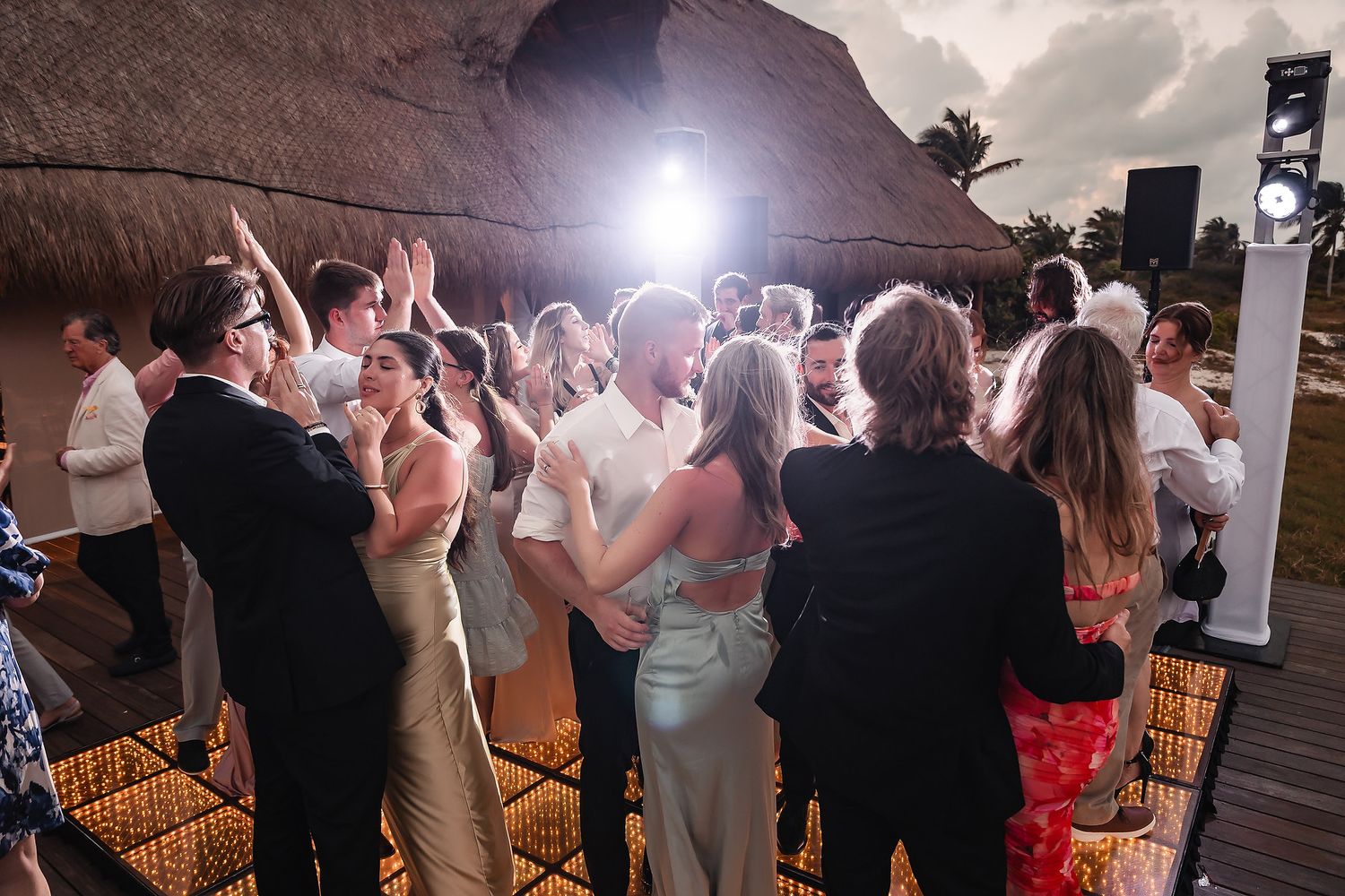 Guests dancing together at an outdoor tropical wedding reception under a thatched roof with bright stage lighting.