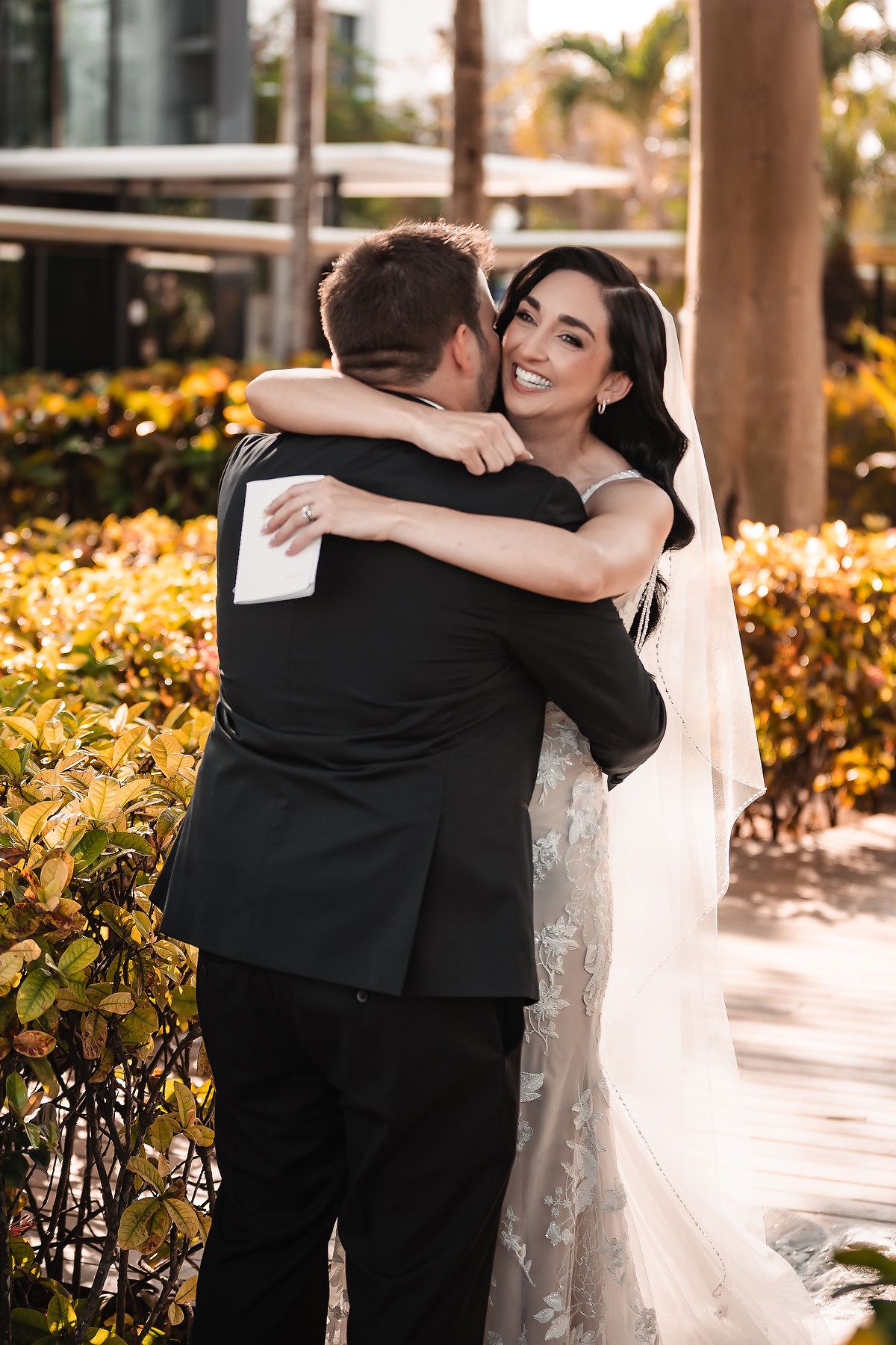 Smiling bride in lace gown and veil hugs groom in black suit outdoors at a wedding.