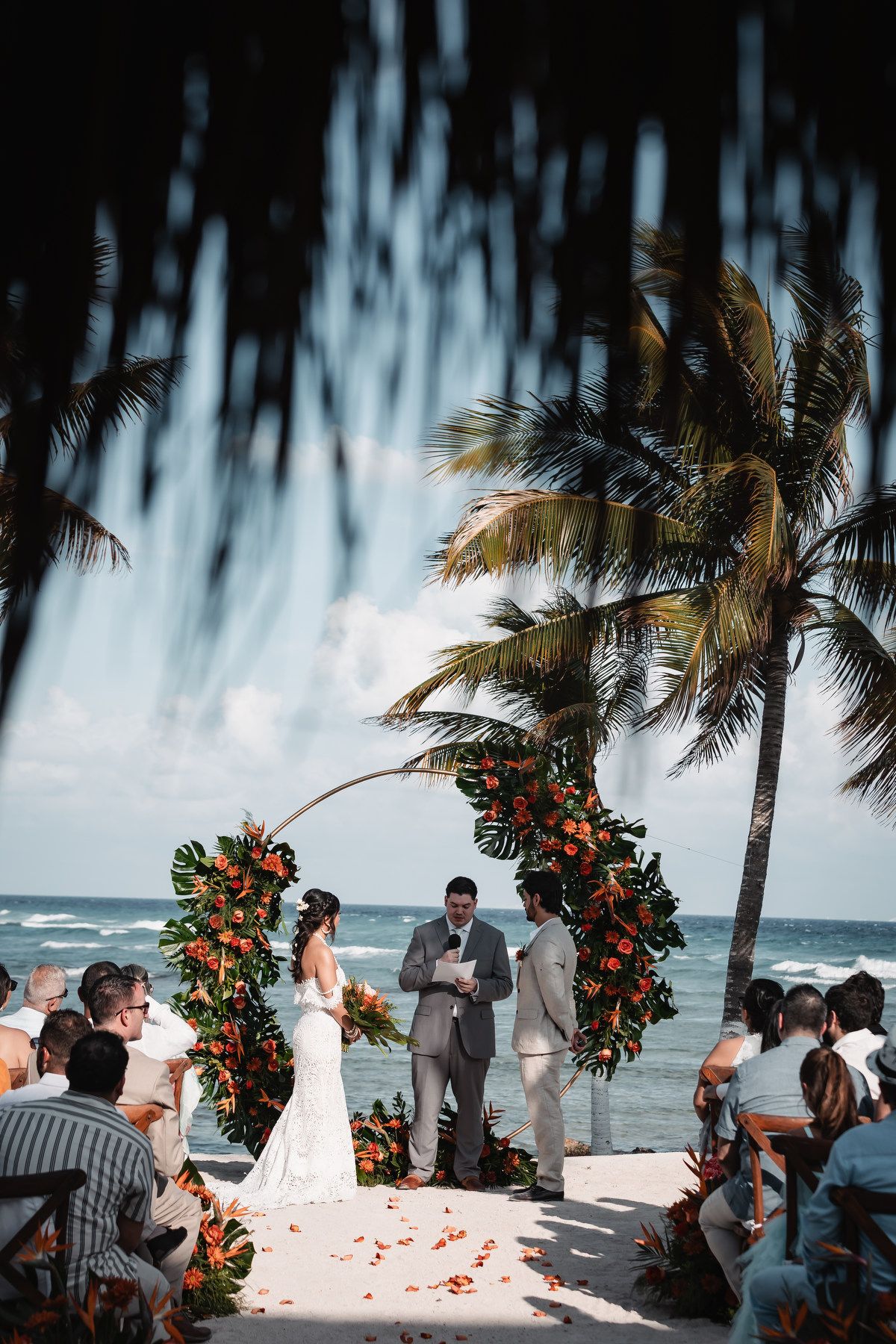 Bride and groom exchange vows at a tropical beach wedding ceremony with floral arch and palm trees.