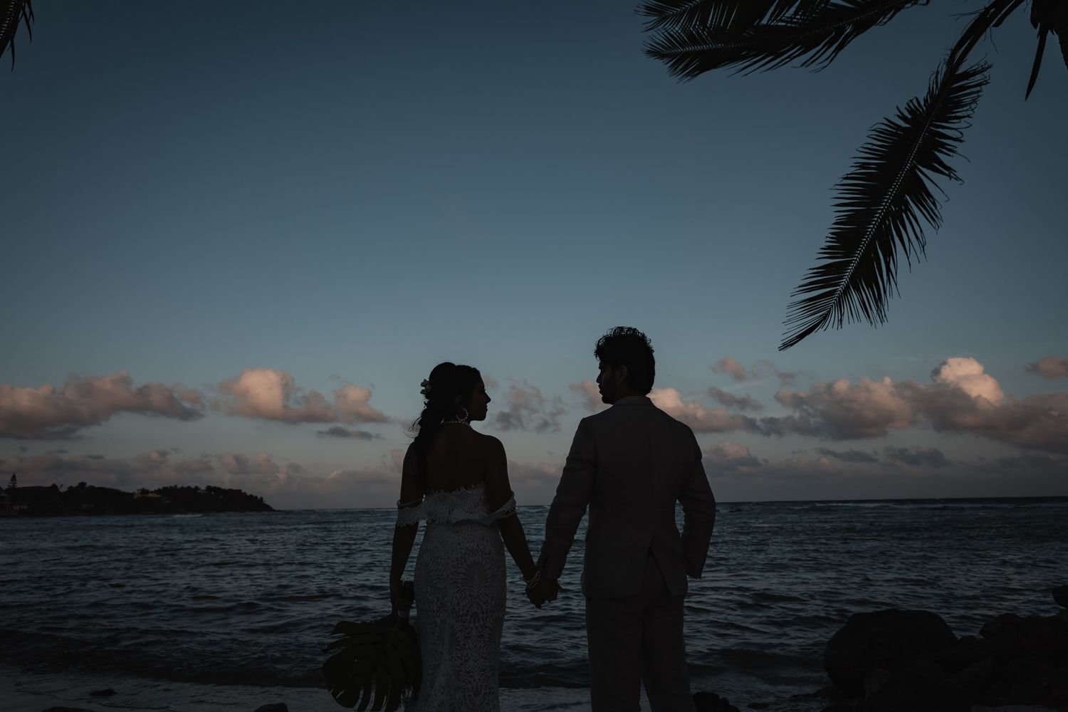 Silhouette of bride and groom holding hands at dusk on tropical beach with ocean and cloudy sky.