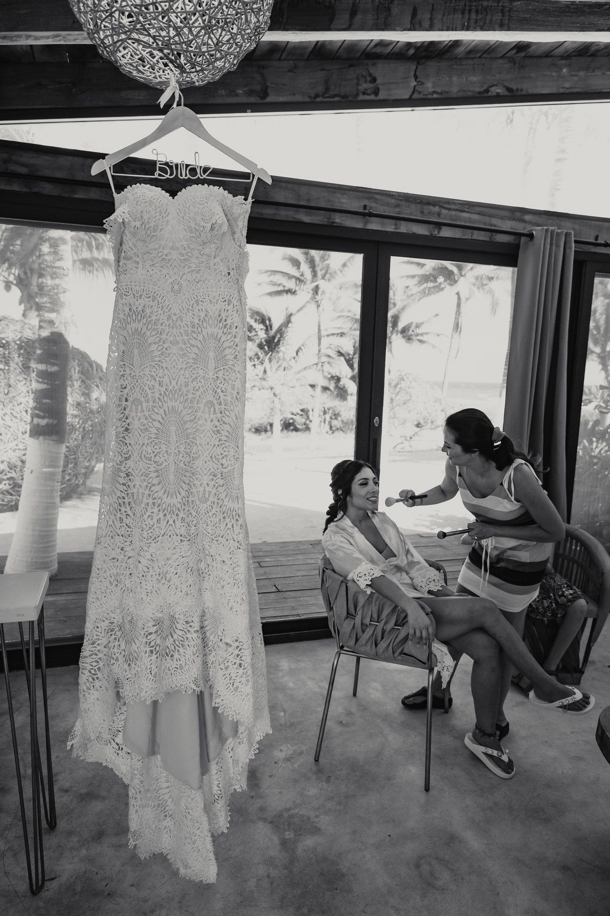 Black and white photo of a lace wedding dress hanging while bride gets ready near tropical windows.