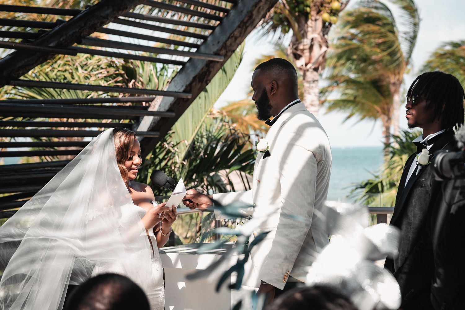 Couple exchanging vows under rustic pergola with palm trees and ocean backdrop.