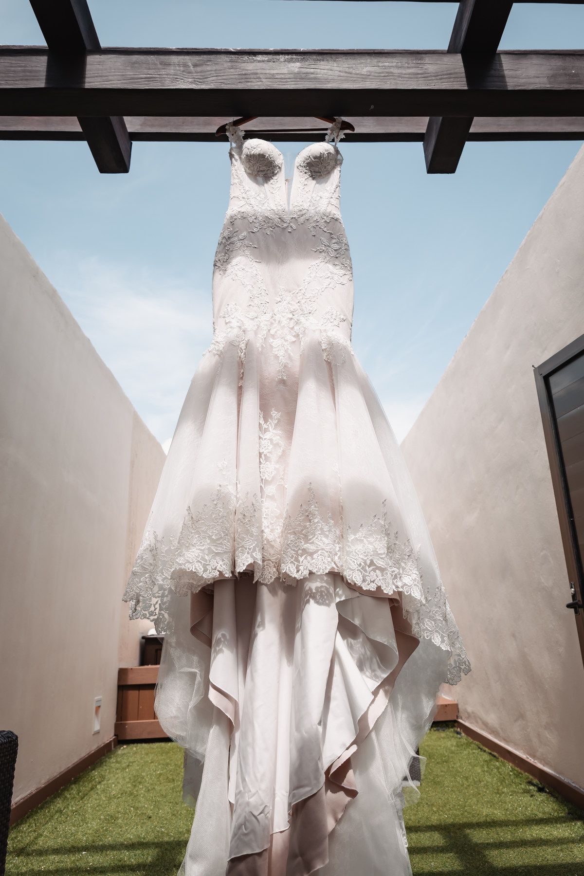Elegant lace mermaid wedding dress hanging outdoors under a pergola against a blue sky.