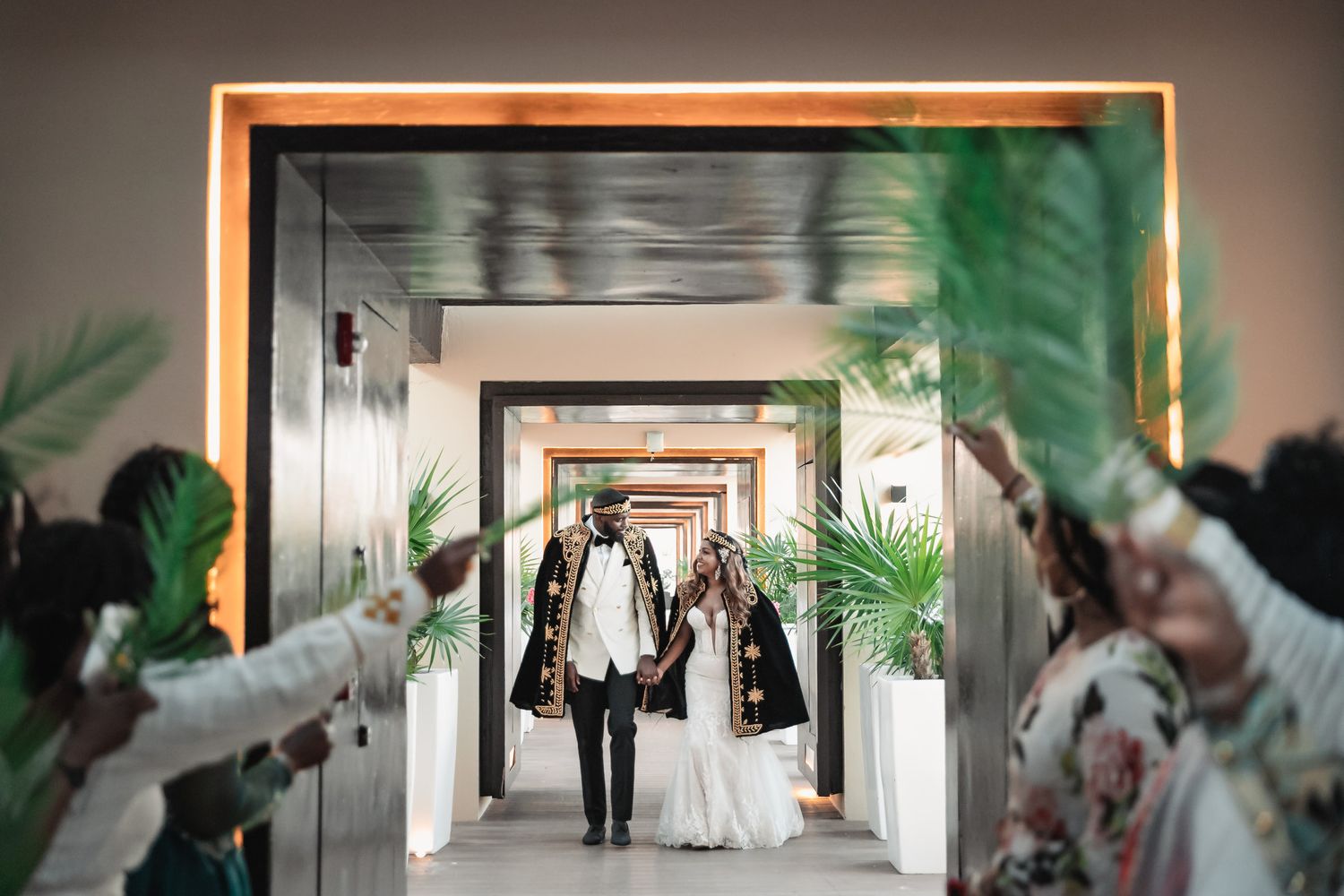 Couple in matching black and gold traditional Ethiopian wedding capes walking through palm-lined corridor.