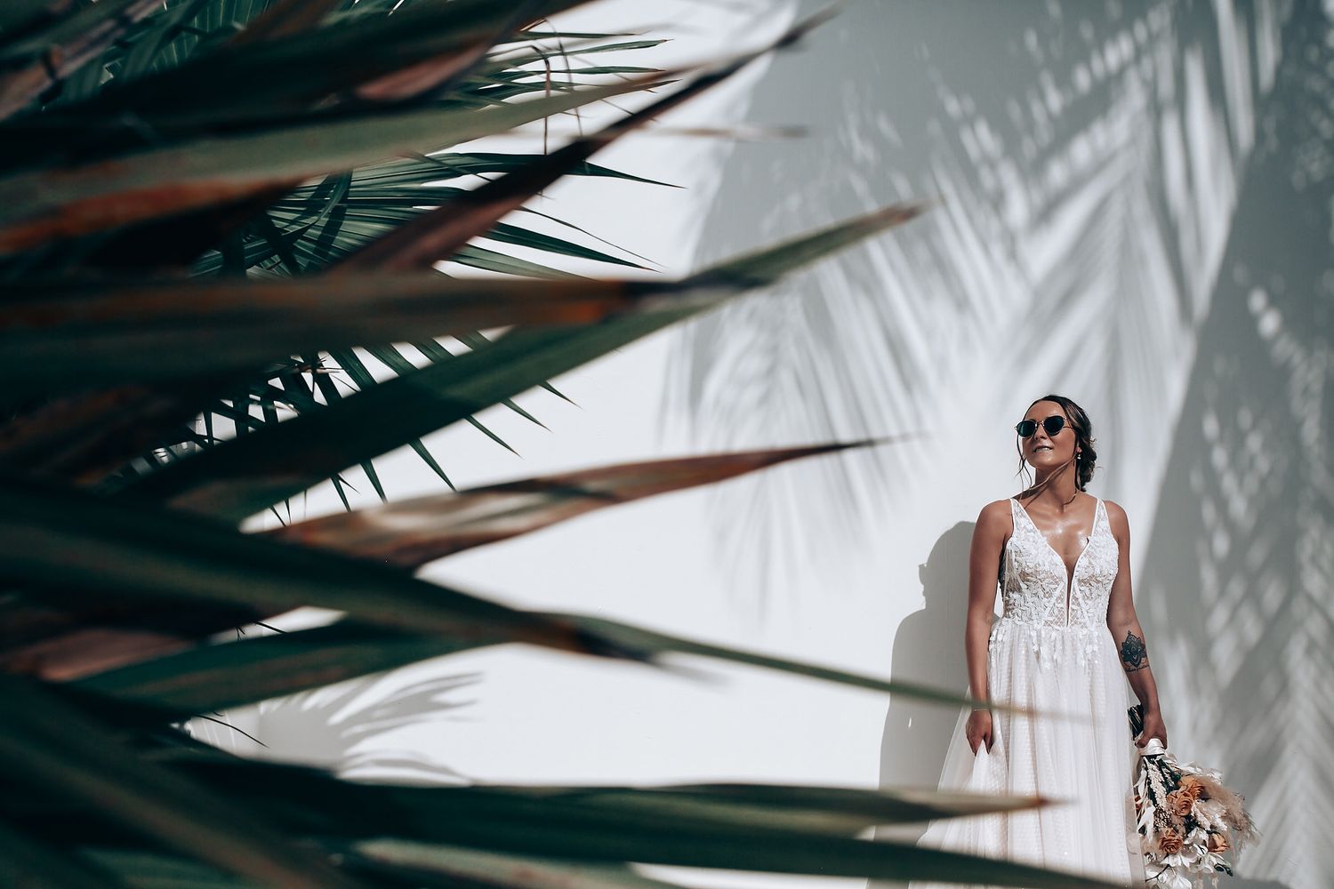 Tropical fashion portrait through palm fronds with dramatic shadow play on a white wall.