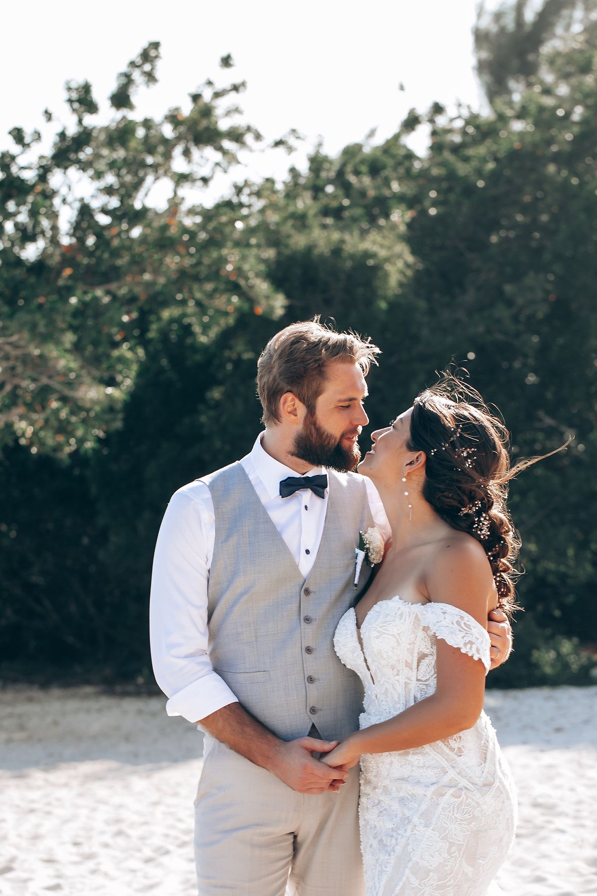 Romantic beach wedding couple embracing and kissing in white dress and beige vest against tropical greenery background.