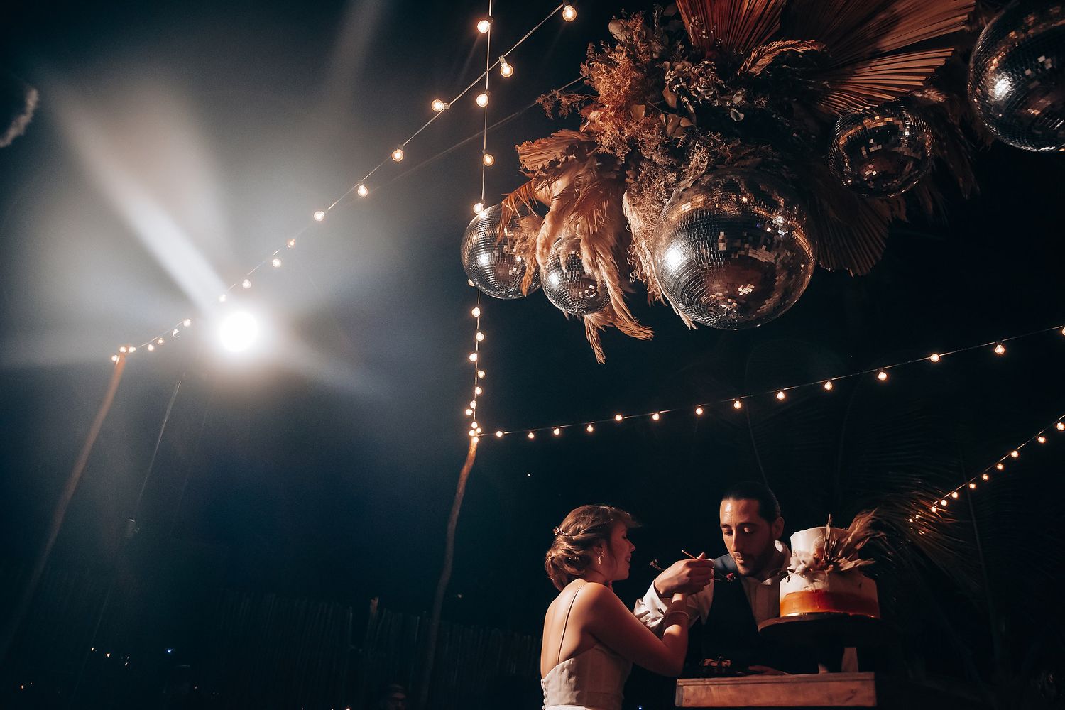 A couple dances their first dance at a wedding reception under warm string lights and disco balls.