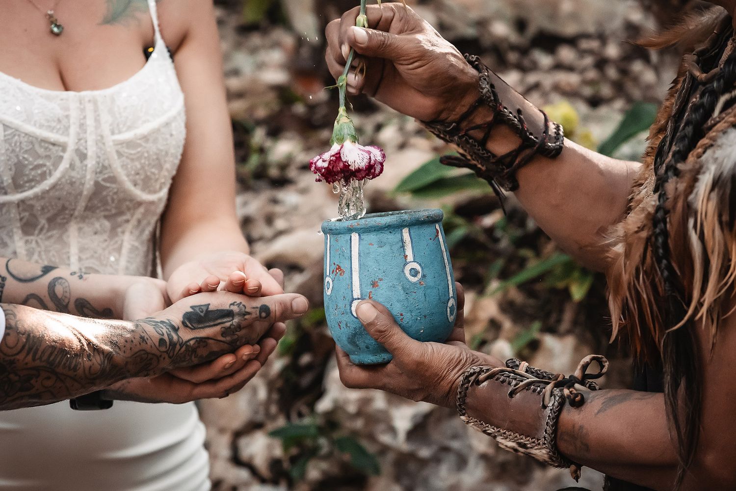 Tattooed hands exchange flowers over a turquoise ceramic cup in a spiritual ritual ceremony outdoors.