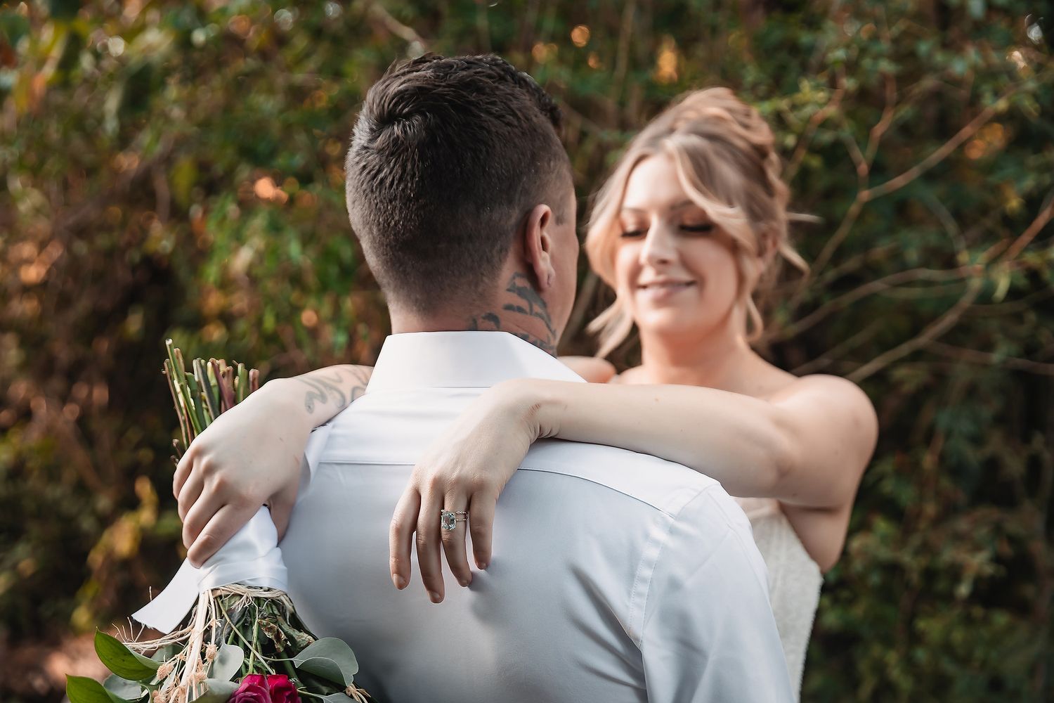 Bride and groom embrace outdoors, woman smiling with arms around mans neck, holding floral bouquet.