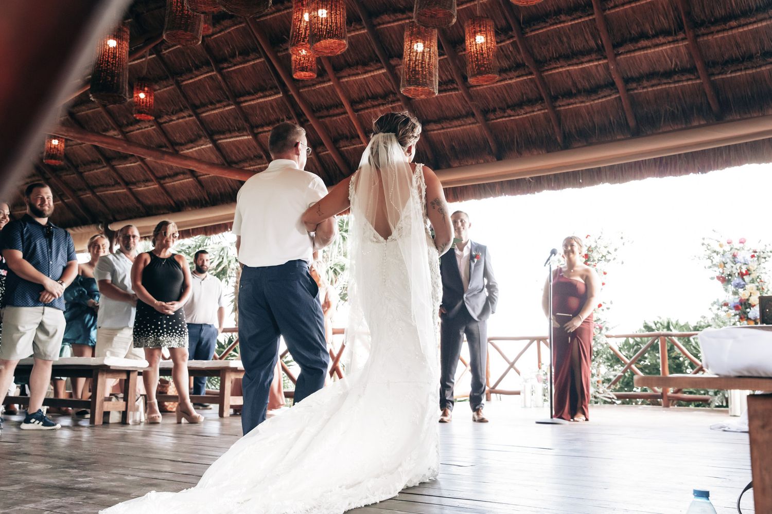 Bride walking down the aisle in a white gown and veil at an outdoor tropical wedding ceremony.