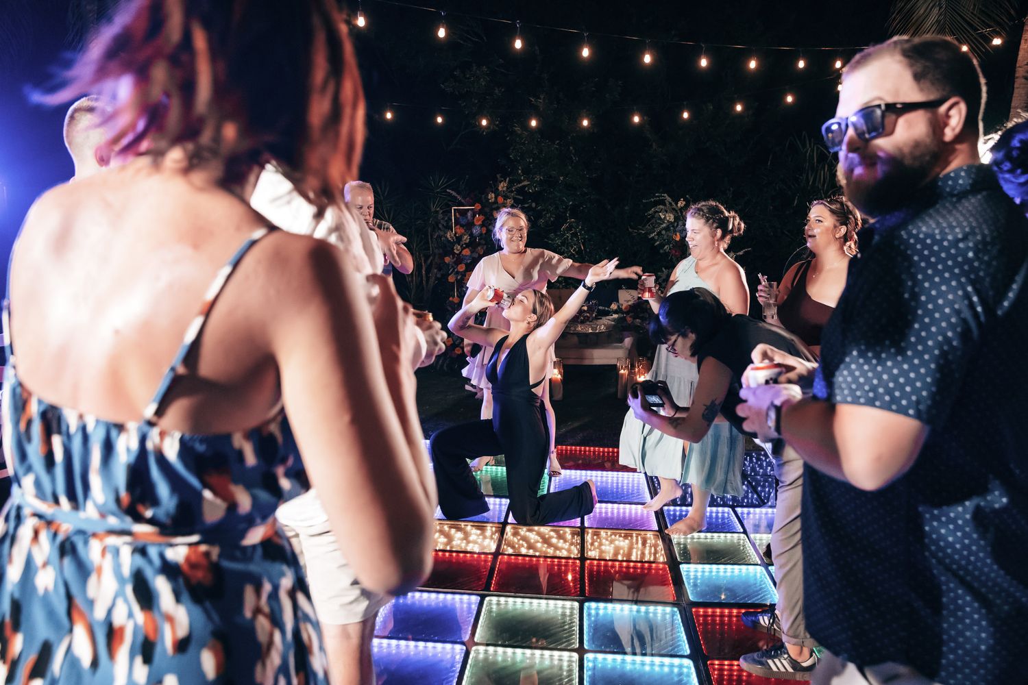 Wedding guests dance and celebrate on a colorful LED dance floor under string lights at an outdoor reception.
