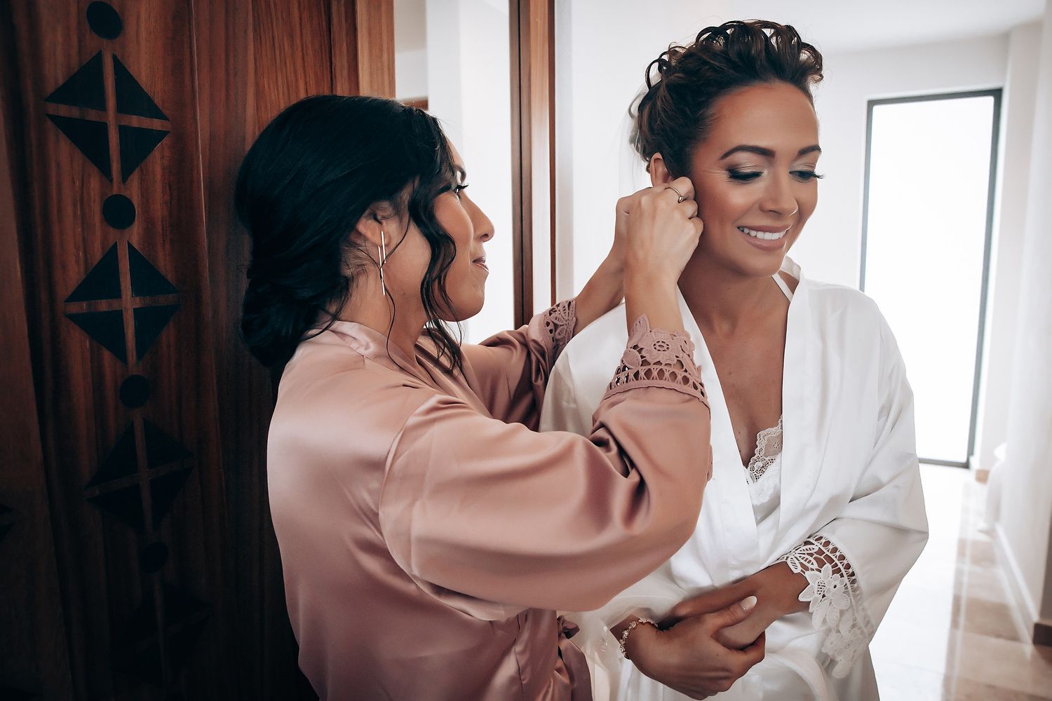 A tender moment during wedding preparations as a bride gets help with her white dress in a sunlit room.