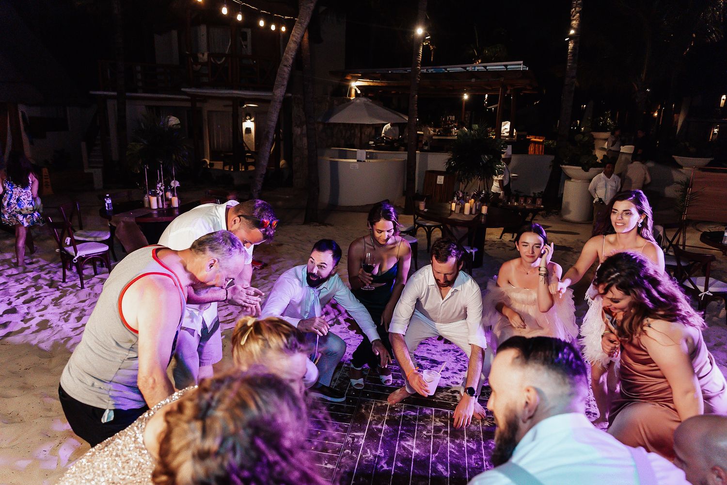Wedding guests gather in a circle on the beach under purple lighting during evening celebrations.