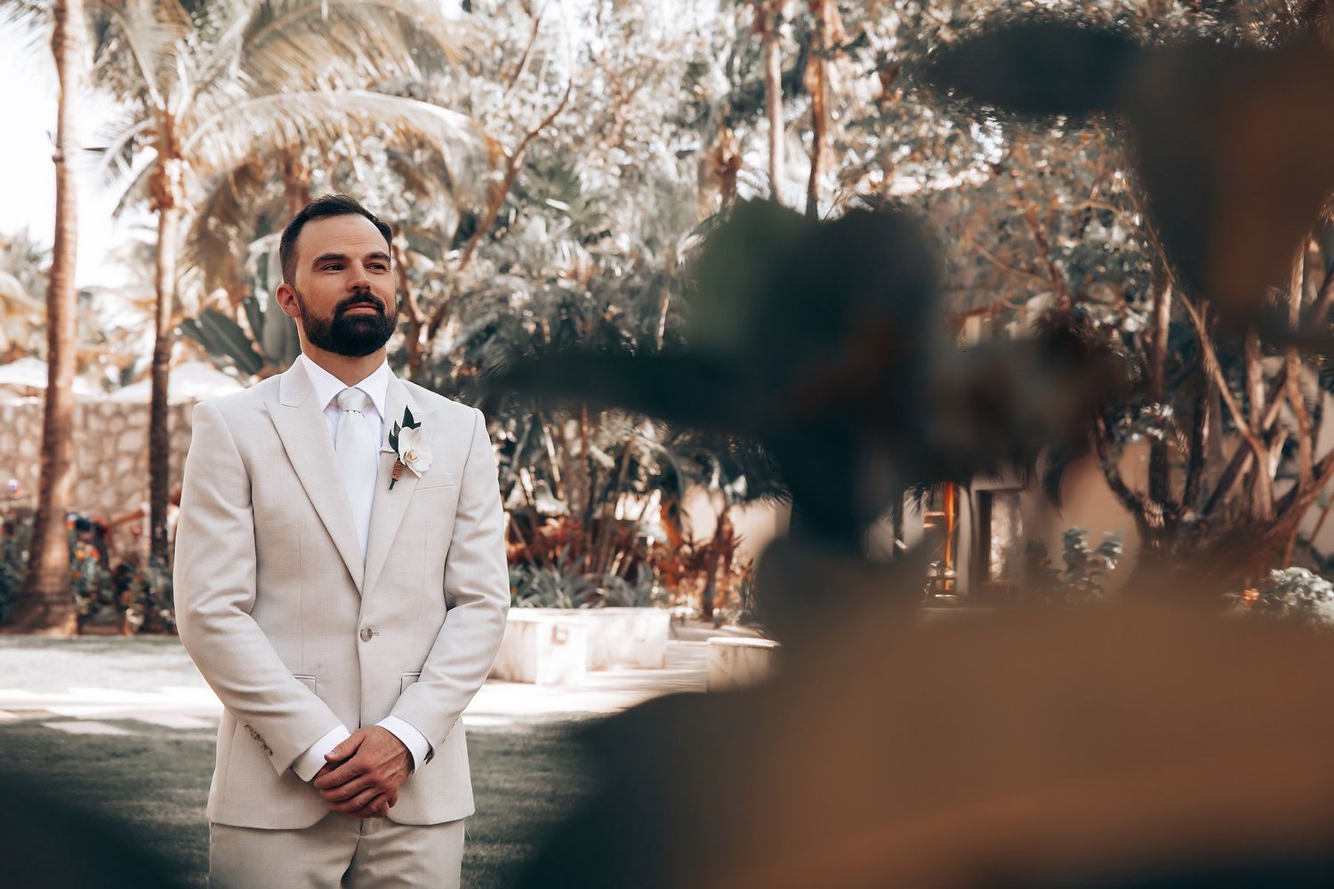 Couple in elegant white wedding attire poses in a sun-dappled garden setting with palm trees and natural lighting.