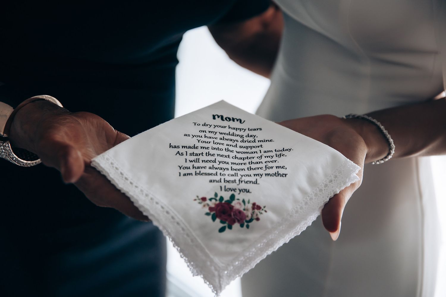A white embroidered handkerchief with sentimental text and floral designs being held up against a light background.