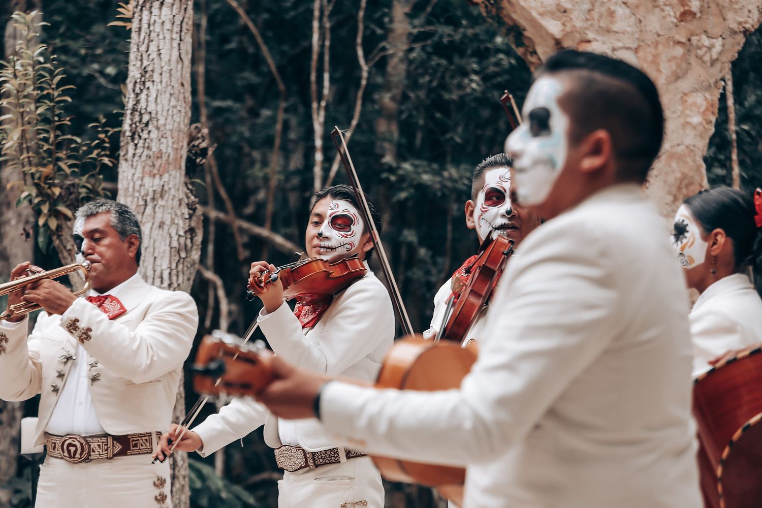 Musicians in white attire perform with brass instruments in an outdoor wooded setting.