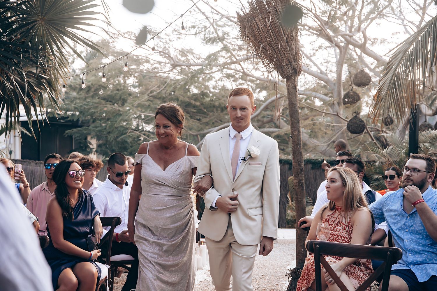 Wedding guests celebrate and dance together at an outdoor reception under palm trees at sunset.
