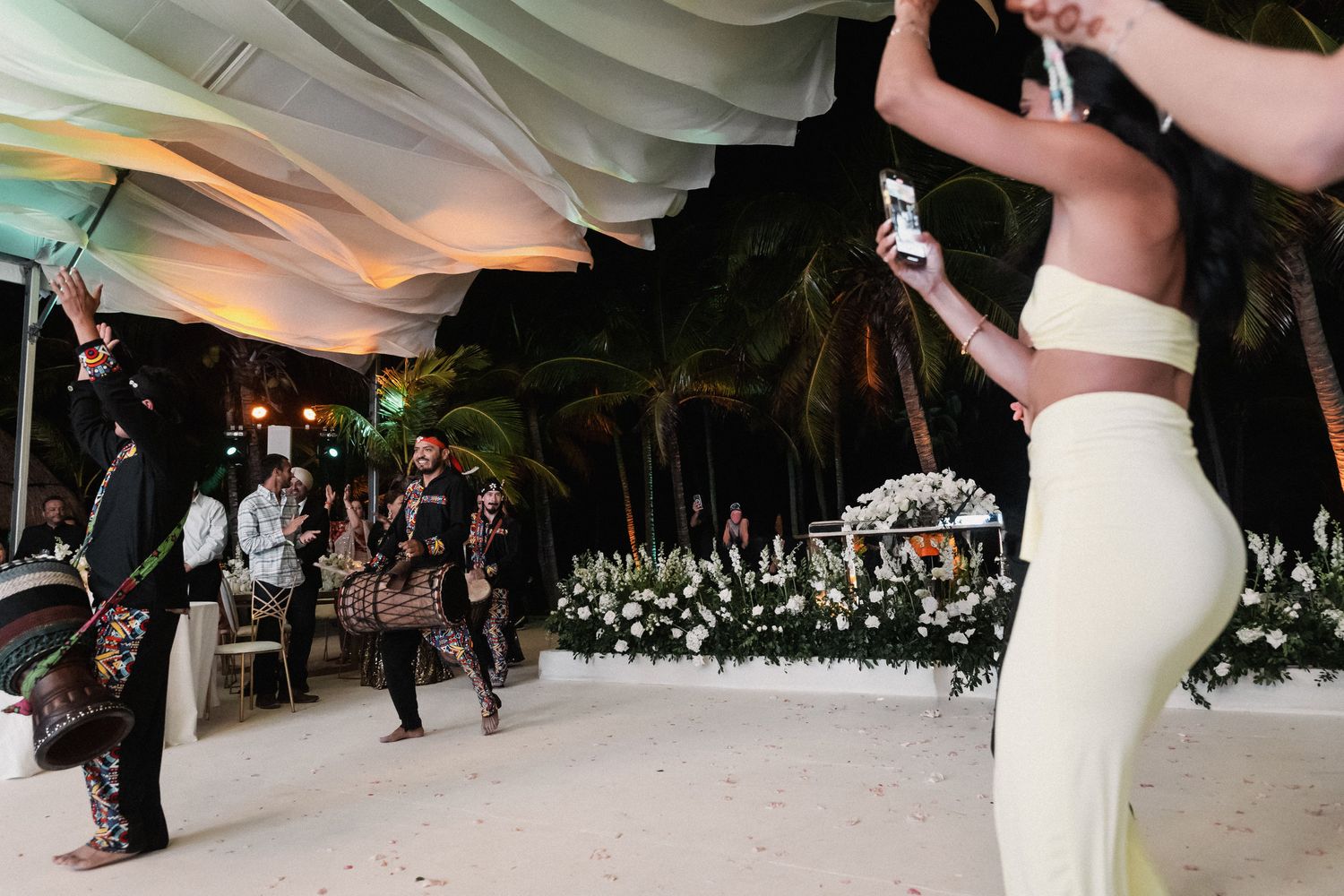 Mariachi band performs for guests dancing under white draped tent with tropical palm trees at beach wedding.