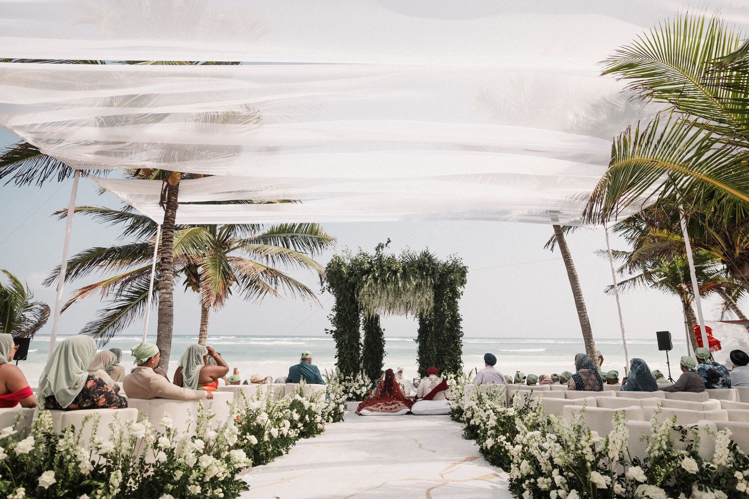 Beach wedding ceremony in progress with guests seated along white floral aisle under draped canopy with greenery arch and ocean backdrop.