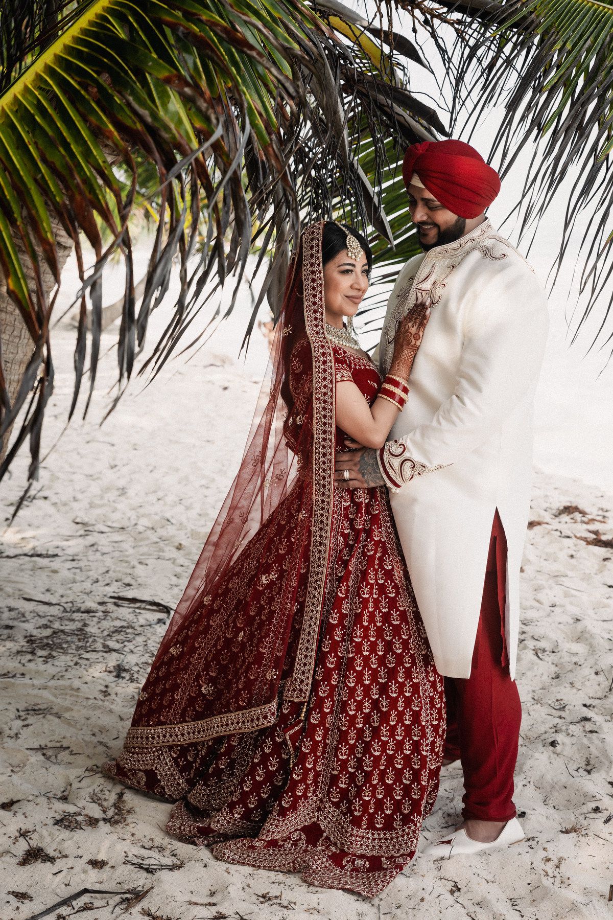 Indian bride in ornate red lehenga and groom in white sherwani embracing romantically under tropical palm tree on sandy beach.