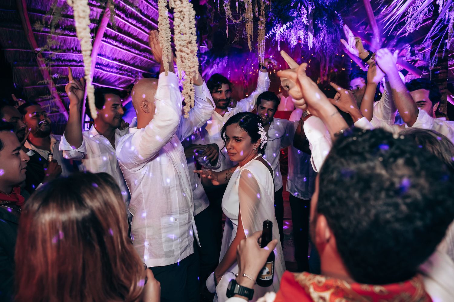 Wedding party dancing under purple lighting at tropical reception venue.