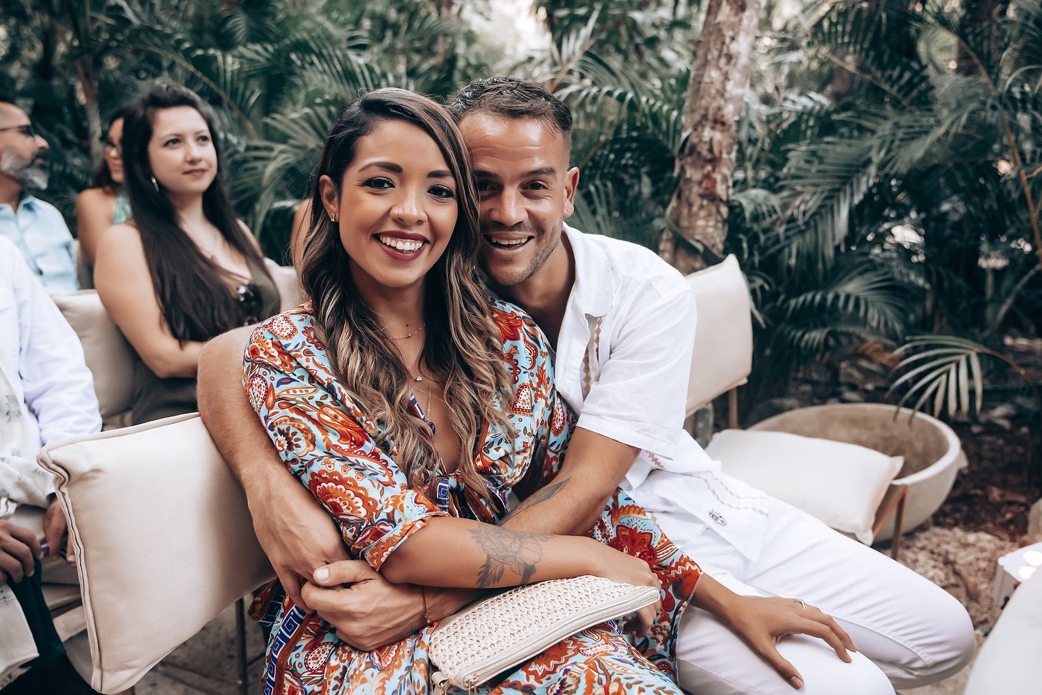 Couple sharing intimate moments on white outdoor furniture surrounded by tropical foliage.
