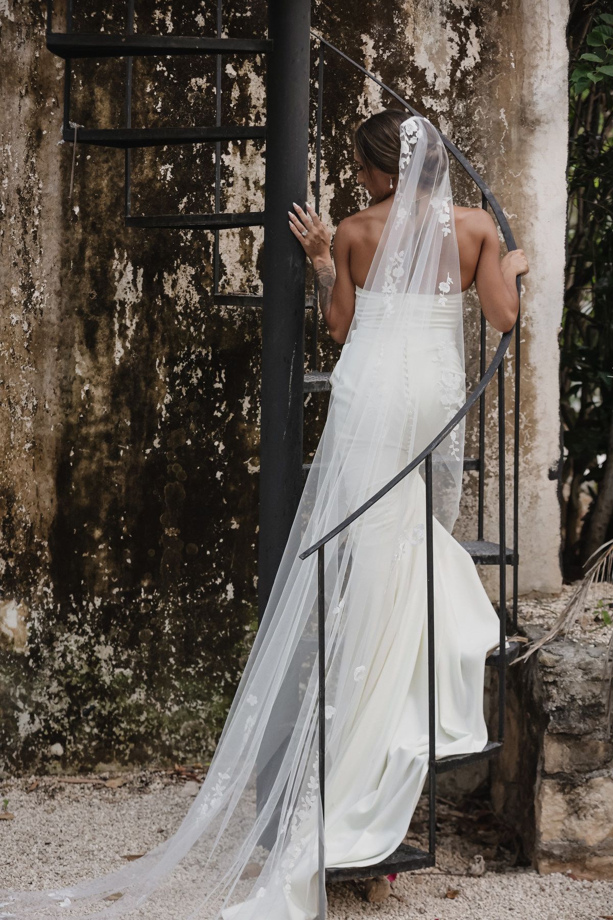 Bride in elegant white gown and lace veil ascending iron staircase against rustic stone wall.
