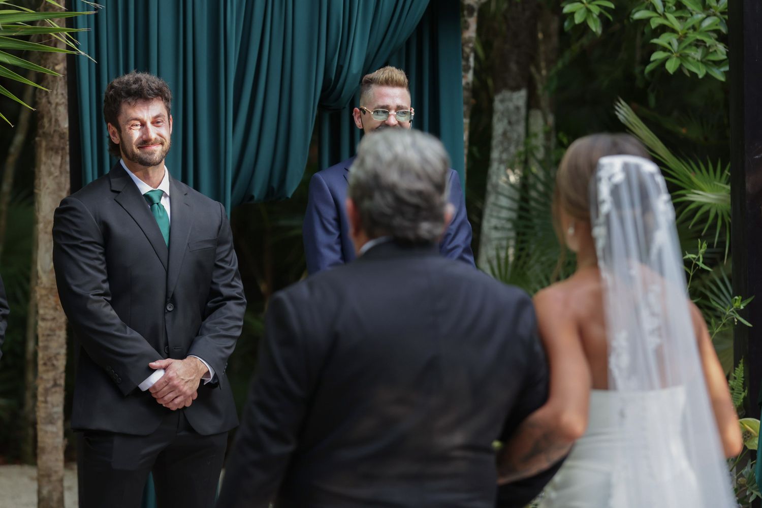 Groom smiles warmly as bride approaches the altar during outdoor wedding ceremony with green draped backdrop.
