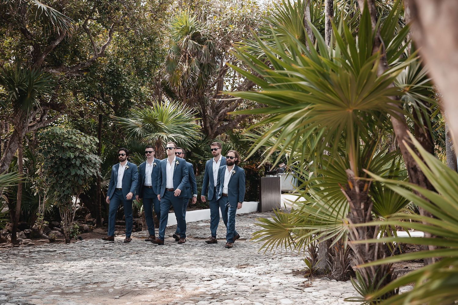 Groomsmen in navy suits walking together along a sandy path surrounded by lush tropical palm trees.