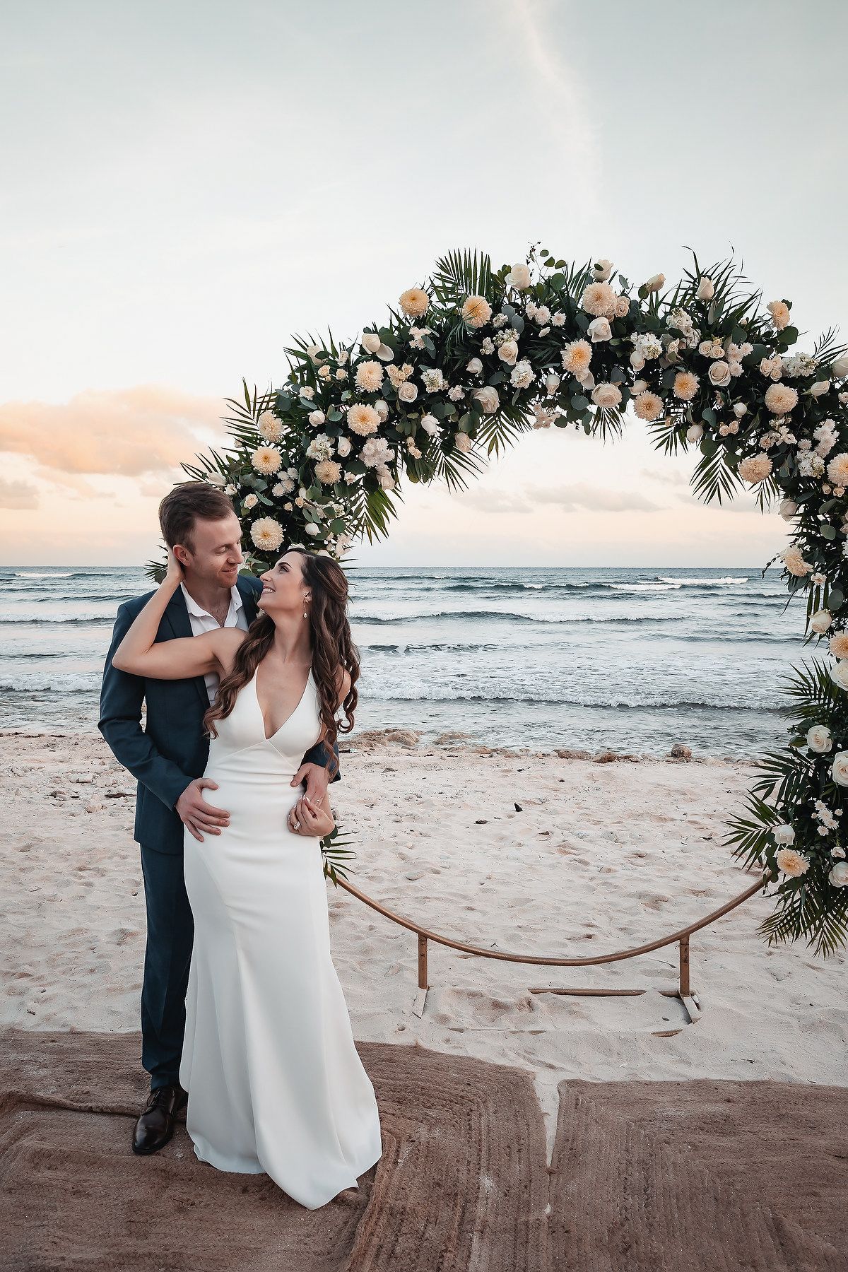 Couple embracing under a floral arch on a beach at sunset, bride in white gown, groom in navy suit.