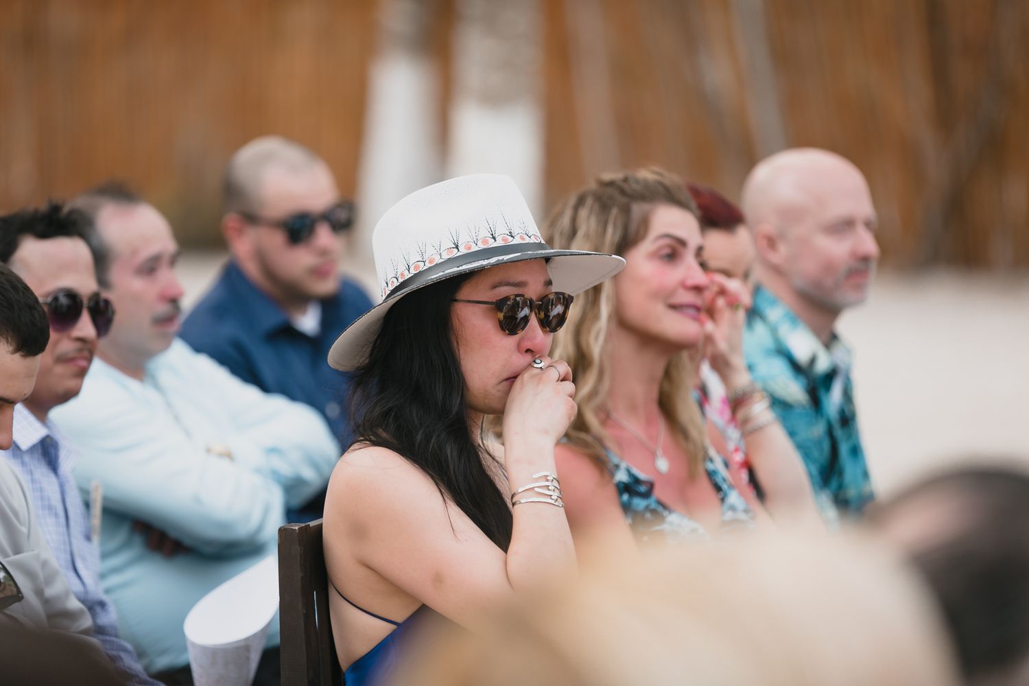 Emotional wedding guest wearing a white hat and sunglasses watches a beach ceremony.